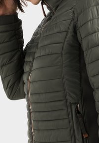 Vadderad grön puffjacka med hög krage, med en hel dragkedja och kontrasterande sidopaneler. Accentuerad med bruna dragkedjedetaljer.