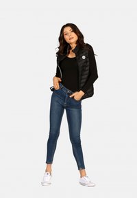 Doudoune noire sans manches avec fermeture éclair et patch logo, associée à un jean bleu ajusté et des baskets blanches. Texture lisse et design slim.