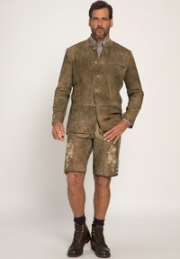 Bruine suède jas met twee knopen en een enkele zak, gecombineerd met bijpassende shorts met bloemenborduursel. Bruine leren laarzen maken de look af.