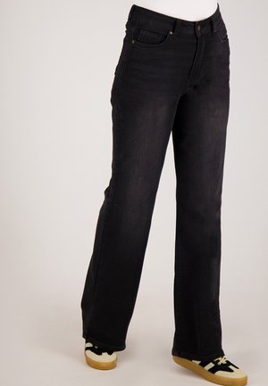 Persoon draagt zwarte recht gesneden jeans met hoge taille en zwart-witte sneakers met rubberen zolen tegen een effen achtergrond.