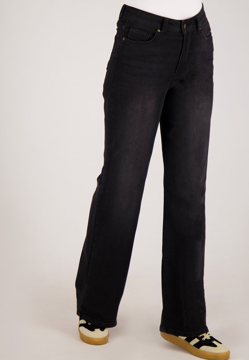 Persoon draagt zwarte recht gesneden jeans met hoge taille en zwart-witte sneakers met rubberen zolen tegen een effen achtergrond.