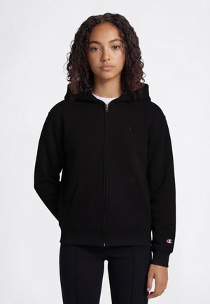 Jonge vrouw met krullend haar, gekleed in een zwarte hoodie met ritssluiting en een zwarte broek, staande tegen een effen witte achtergrond.