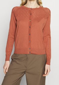 Svetr v rzi s texturovaným vzorovaným průramkem a zapínáním na knoflík. Má dlouhé rukávy a žebrované manžety a lem.