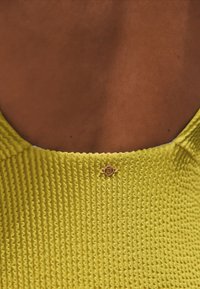 Vêtement jaune texturé avec un motif côtelé, comportant un petit accent circulaire en métal doré au niveau de l'encolure arrière.