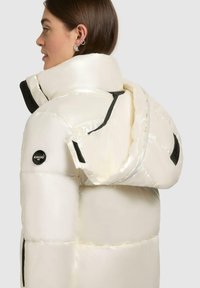 Puffer jacket in glossy cream fabric with a high collar, oversized hood, black accents, and logo detail on the sleeve.