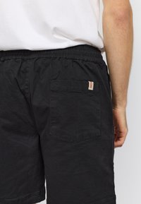 Achteraanzicht van een persoon die zwarte shorts met elastische taille en achterzak draagt, gecombineerd met een wit t-shirt dat is ingestopt, arm langs het lichaam.