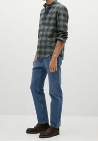Grünes und graues karierter Hemd, zugeknöpft mit einer Brusttasche, blaue Jeans und dunkelbraune Schnürschuhe, stehend vor einem hellen Hintergrund.
