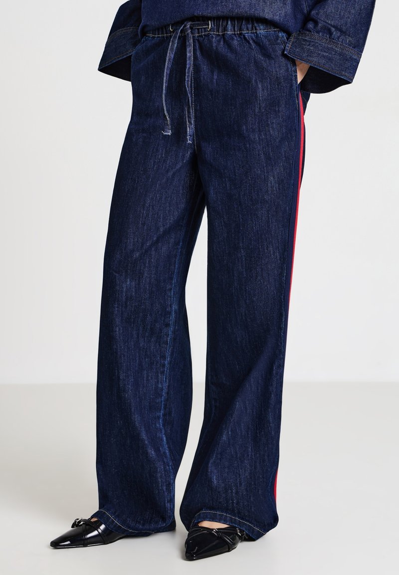 Pantaloni in denim scuro a gamba larga con vita elastica, cordino e strisce laterali rosse, abbinati a scarpe nere con punta.