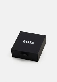 Cutie rectangulară neagră cu un finisaj mat, având un logo „BOSS” argintiu pe față. Include un tab pentru deschidere.