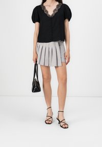 Blouse noire avec un col en dentelle et des manches bouffantes, associée à une jupe grise plissée et des sandales noires à lanières. Tenant un petit sac à main noir.