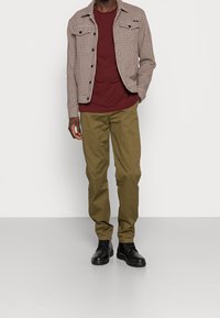 Giacca marrone a scacchi houndstooth con due tasche sul petto, una t-shirt bordeaux, pantaloni verde oliva e stivali neri con lacci. Design classico e strutturato.