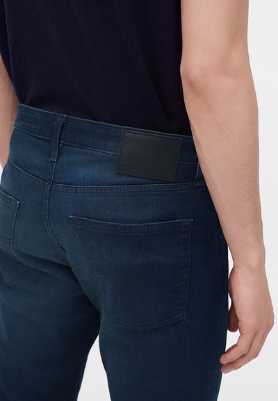 Personne portant un jean bleu foncé et une chemise noire, vue de dos avec un focus sur la poche et la ceinture.