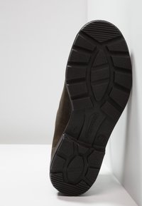 Bota de ante marrón con una suela de goma negra resistente que presenta un dibujo de patrón y la marca "Blundstone", vista desde la parte inferior.