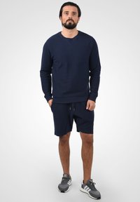 Sweatshirt marine en tissu texturé, col rond et poignets côtelés, associé à un short marine assorti et des chaussures de sport grises.