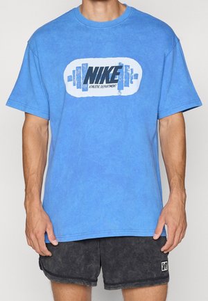 Homme portant un t-shirt Nike bleu avec un logo haltère et un short de sport noir, debout droit devant un fond clair uni.