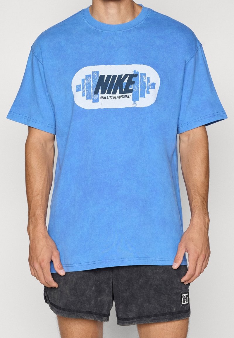 Homme portant un t-shirt Nike bleu avec un logo haltère et un short de sport noir, debout droit devant un fond clair uni.