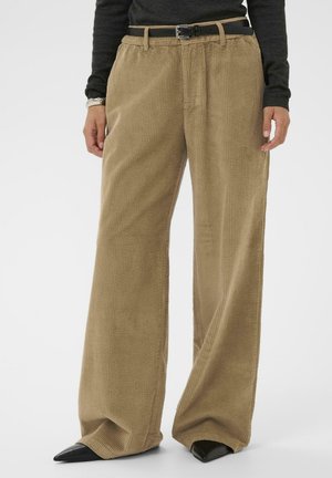 Femme portant un pantalon en velours côtelé beige à jambes larges, des chaussures noires à bout pointu, une ceinture noire et un haut à manches longues gris foncé, se tenant devant un fond blanc.