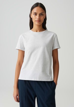 Giovane donna con capelli lunghi e scuri, che indossa una maglietta a maniche corte di colore grigio chiaro e pantaloni scuri, in piedi contro uno sfondo chiaro.