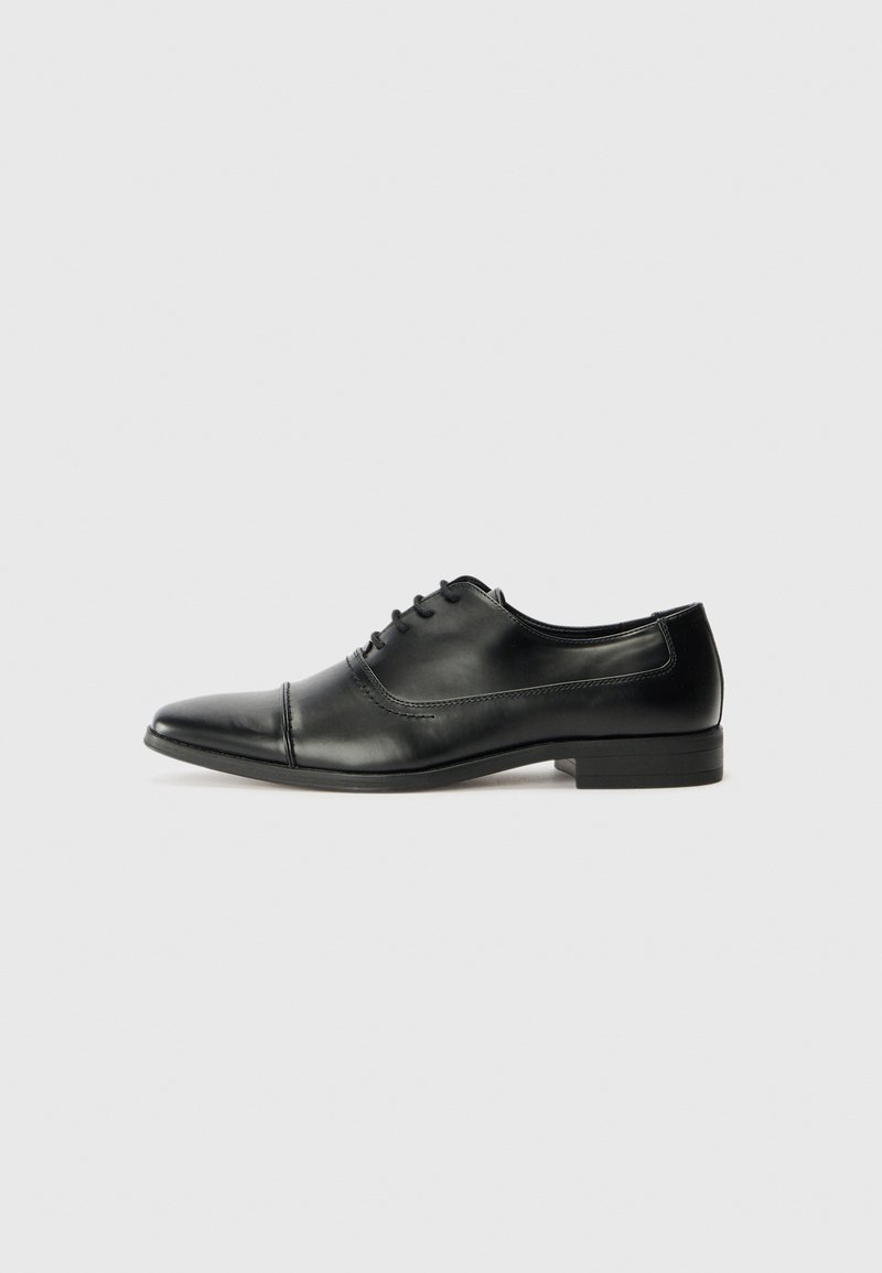 Chaussure Oxford en cuir noir pour homme avec lacets et petit talon, présentée de profil sur un fond gris clair uni.