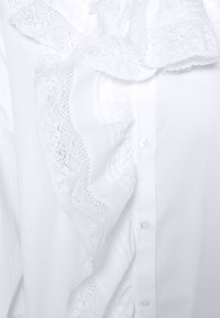 Witte blouse met verticale ruches en fijne kanten details langs de kraag en de knoopsluiting aan de voorkant.