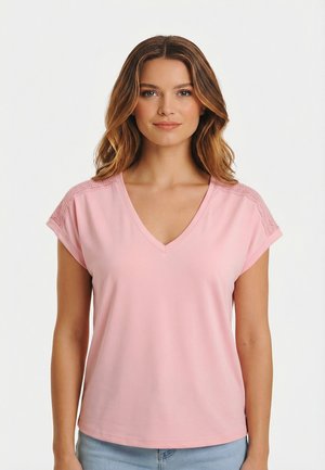 Vrouw met lichtbruin golvend haar, gekleed in een lichtroze V-hals T-shirt met korte mouwen en kanten details op de schouders, en lichtblauwe spijkerbroek.