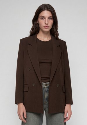 Junge Frau mit langem dunklem Haar trägt einen dunkelbraunen zweireihigen Blazer über einem passenden Oberteil und grauen Jeans und steht vor einem schlichten Hintergrund.