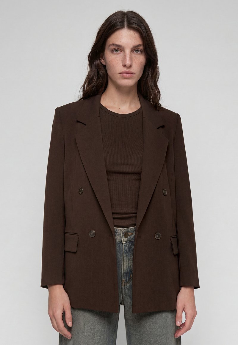 Young woman with long dark hair wears a dark brown double-breasted blazer over a matching top and gray jeans, standing against a plain background.
