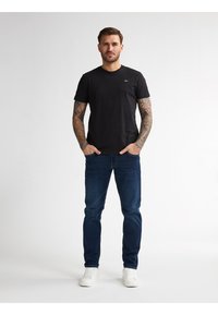 Černé bavlněné tričko s krátkým rukávem kombinované s tmavě modrými džíny. Model má na sobě bílé tenisky; tetování viditelné na pažích. Jednoduchý, ležérní vzhled.