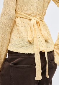 Gul teksturert langermet topp med et stoffbelte knytt i sløyfe i midjen, kombinert med mørkebrune bukser med store baklommer.