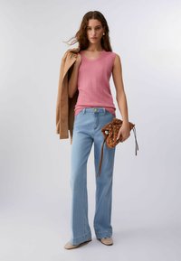 Femme portant un haut rose sans manches, un jean évasé bleu clair, tenant une pochette marron cloutée et une veste beige sur l'épaule.
