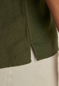 Camicia di lino verde scuro con uno spacco laterale all'orlo, caratterizzata da una texture leggera e un taglio dritto, abbinata a un tessuto bianco sporco al di sotto.
