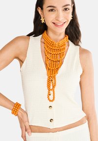 Hvid tekstureret ærmeløs top med V-hals og gyldne knapper, parret med en orange perlekæde og matchende armbånd.