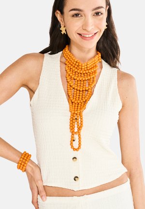 Hvid tekstureret ærmeløs top med V-hals og gyldne knapper, parret med en orange perlekæde og matchende armbånd.