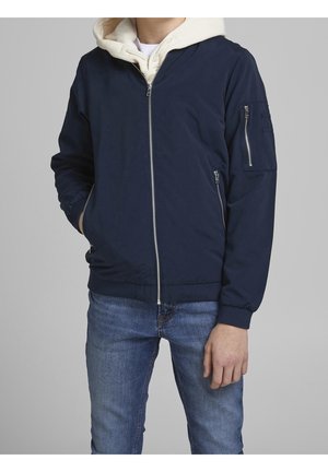 Homme portant une veste bomber bleu marine avec des poches zippées à l'avant et aux manches par-dessus un sweat à capuche beige clair et un jean bleu, debout devant un fond gris uni.