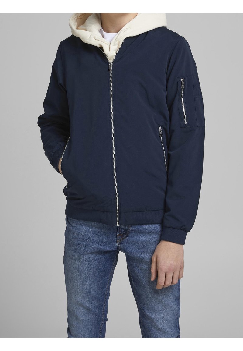 Homme portant une veste bomber bleu marine avec des poches zippées à l'avant et aux manches par-dessus un sweat à capuche beige clair et un jean bleu, debout devant un fond gris uni.