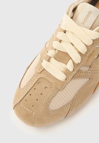 Lysebrun ruskindsneaker med meshpaneler, cremefarvede snørebånd og perforerede detaljer. Har diskret brandlogo i guld. Rundet tådesign.