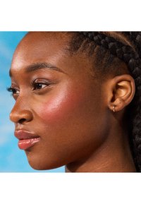 Vista de perfil de una persona de piel oscura con el cabello trenzado, que lleva pequeños pendientes de aro de oro. La piel tiene un brillo natural; un rubor rosado añade color.