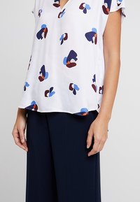 Blusa bianca a maniche corte con scollatura a V, caratterizzata da motivi astratti in blu, rosso e marrone. Tessuto leggero, vestibilità rilassata.