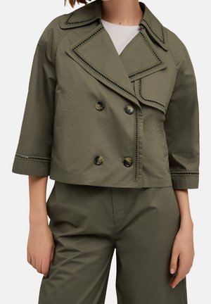 Mujer con chaqueta corta de doble botonadura verde oliva con ribete decorativo y pantalones a juego de talle alto sobre un fondo liso.