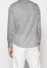 Pitkähihaisessa paidassa on valkoista ja harmaata väriä, jossa on geometrinen kuvio. Siinä on kaulus, napitettavat hihansuut ja kaareva helma. Materiaalina on puuvillasekoitus.