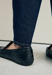 Zwarte leren moccasins met een krokodillenstructuur, lage profield en flexibele zool, gecombineerd met donkere denim jeans met een omgekrulde zoom.