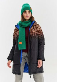 Žena v leopardom vzorovanom a čiernom prešívanom kabáte, zelenej pletenej čiapke a šatke, vínovom topu a svetlo šedých džínsoch na bielom pozadí.