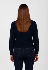 Maglione lavorato a maglia blu navy con polsini e orlo a coste, dal taglio classico. Indossato con jeans in denim scuro che mostrano cuciture visibili.