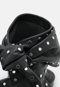 Ceinture en cuir noir avec des clous en argent nouée, mettant en valeur la texture et les détails métalliques sur un fond blanc.