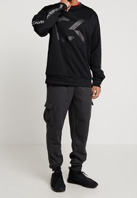 Sweat-shirt noir avec logo, manches longues et col rond ; associé à un pantalon cargo gris foncé avec poches latérales et poignets élastiques.