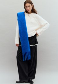 Pulover alb tricotat supradimensionat cu manșete drepte, asortat cu pantaloni largi negri. O eșarfă vibranta albastră cu franjuri cade pe un umăr.