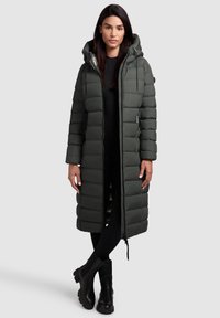 Lange, donkergroene pufferjas met een capuchon, voorzien van horizontale baffles, een voorkleurige rits en zakken. Draagt over zwarte leggings en laarzen.