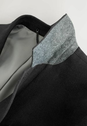 Giacca nera con colletto in feltro grigio, dotata di una fodera interna liscia. Il colletto presenta un bordo cucito per un tocco di dettaglio in più.