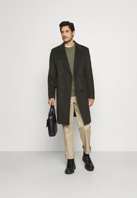 Palton dublu din lână de culoare verde închis, pulover verde deschis, pantaloni bej, geantă din piele neagră și cizme negre cu șiret.