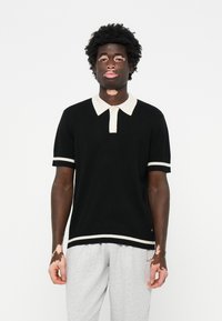 Fekete, rövid ujjú polo ing, krémszínű gallérral és szegélydíszítéssel, puha anyagból készült, szürke melegítőnadrággal párosítva.